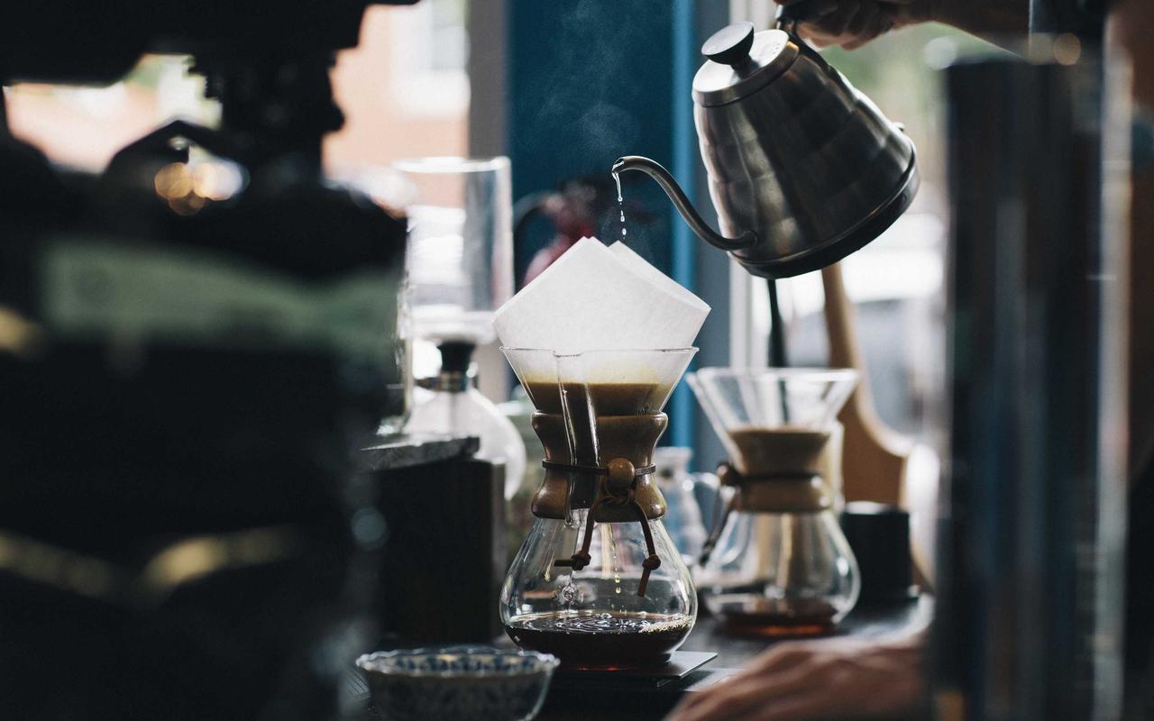 Water poured from a steel kettle into a paper filter over a glass carafe on a dark counter