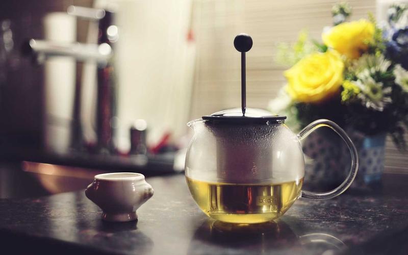 A glass teapot with a steel plunger beside a small white cup and yellow roses