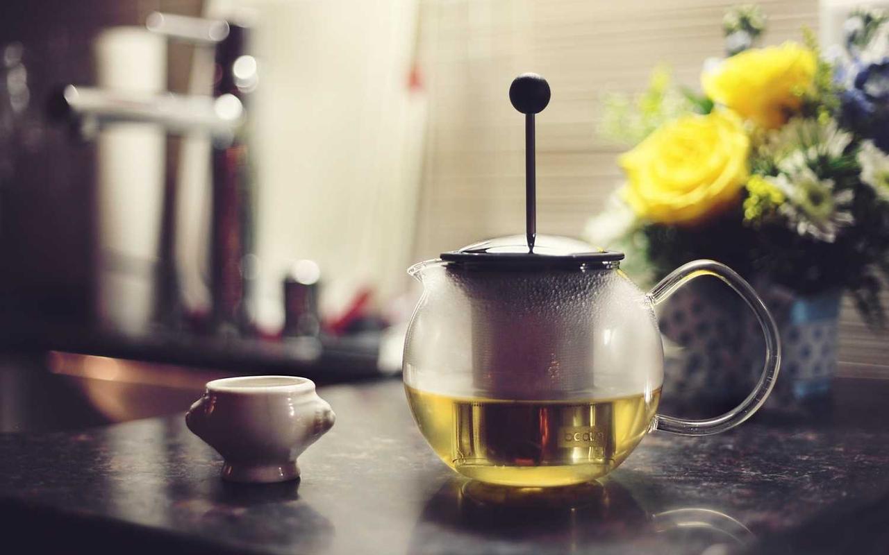 A glass teapot with a steel plunger on a dark counter, a small white cup beside it and yellow roses behind