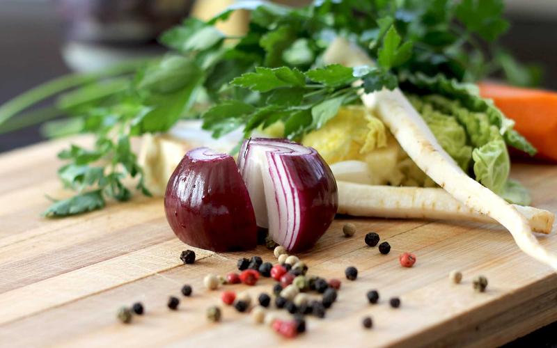 A halved red onion, parsnips, carrots and peppercorns on a wooden chopping board