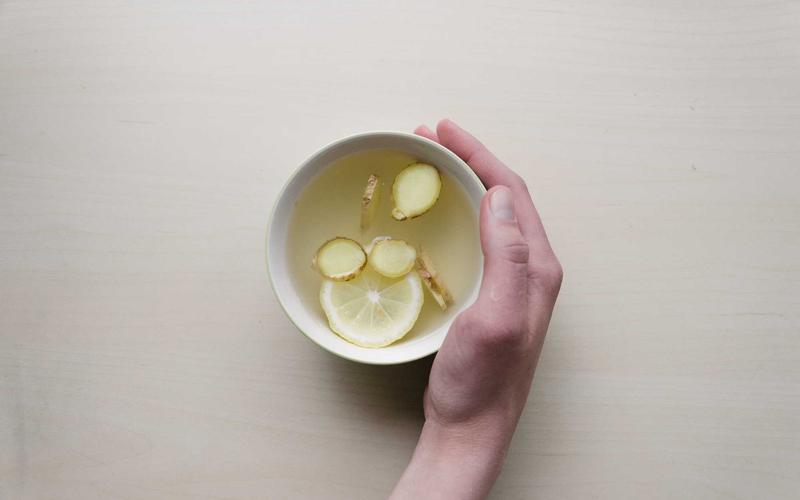 A hand resting beside a white bowl holding slices of lemon and ginger in water, on a pale table