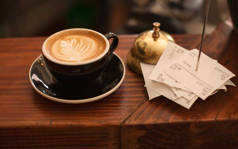 A cappuccino on a dark saucer beside a brass counter bell and a spike of paper receipts